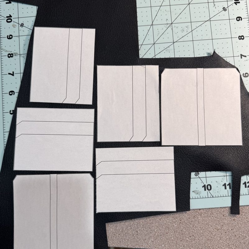 Six paper pattern pieces laid out on a sheet of black leather over a gridded cutting mat