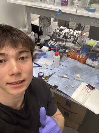 Ben in gloves in the Sonkusale lab, with the bench behind him: a probe station, reagent bottles, tools and finished arrays